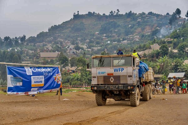 A WFP truck loaded with supplies is parked near a banner reading “Chakula Sokoni” at a distribution site, with hills and houses in the background.